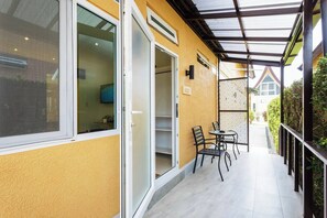 Ein überdachter Außenbereich mit gelber Wand, einer Schiebetür aus Glas, einem kleinen Tisch mit Stühlen und einem Metallzaun.
