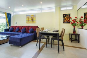 A modern living room with a blue sofa, dining table, and chairs, a painting on the wall, and a vase with red flowers.
