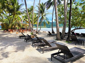 A beachside lounge area with lounge chairs and tables, palm trees, and a view of the sea.