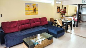 A modern living room with a blue sofa, a glass coffee table, and a dining area with wicker chairs. - Beach Villa 21D Blue Haven Bay Resort - 4 Rooms (Ko Chang)