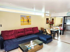 A modern living room with a blue sofa, a glass coffee table, and a dining area with wicker chairs.