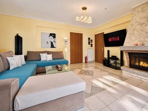 Een woonkamer met een bank, poef, open haard en een flatscreen-tv waarop 'NETFLIX' wordt weergegeven.