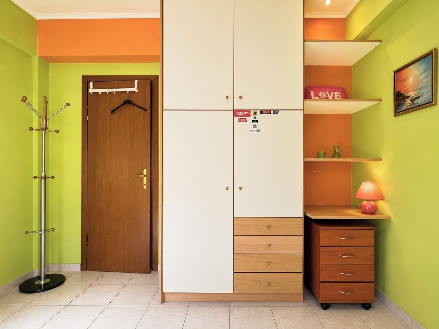 Une pièce avec une porte en bois, un porte-manteau, une armoire avec plusieurs tiroirs, une étagère avec une lampe et un tableau encadré au mur.