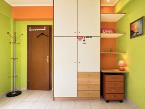 Een kamer met een houten deur, een kapstok, een kledingkast met meerdere lades, een plank met een lamp en een lijst met een afbeelding aan de muur.