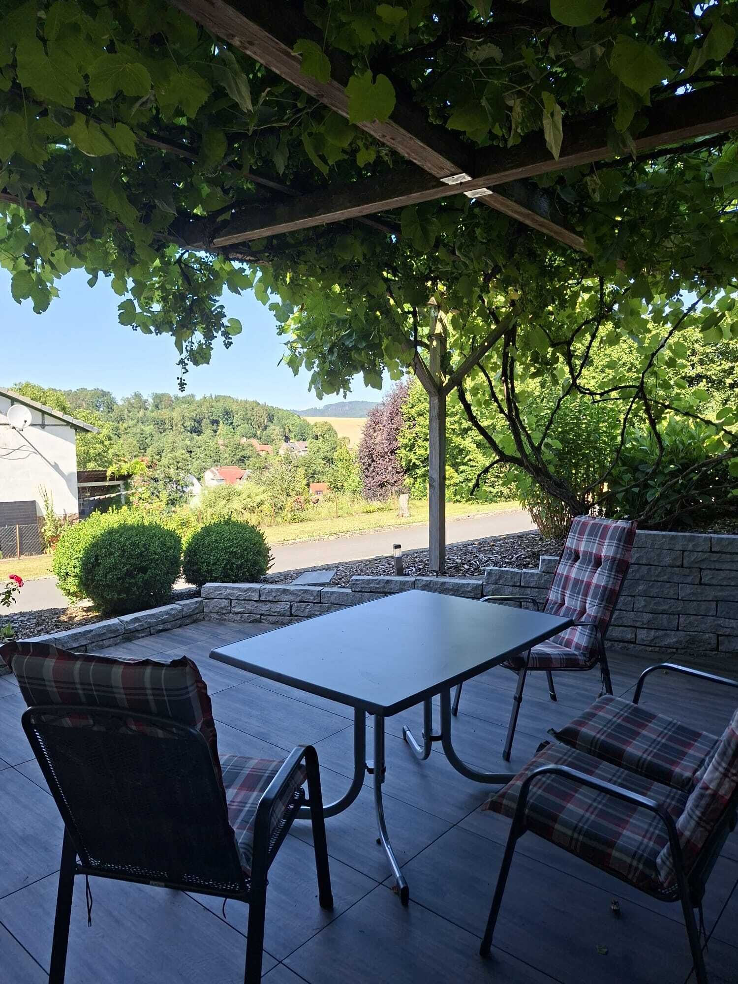 Eine Terrasse im Freien mit Tisch und vier Stühlen, umgeben von viel Grün und Blick auf die Landschaft.