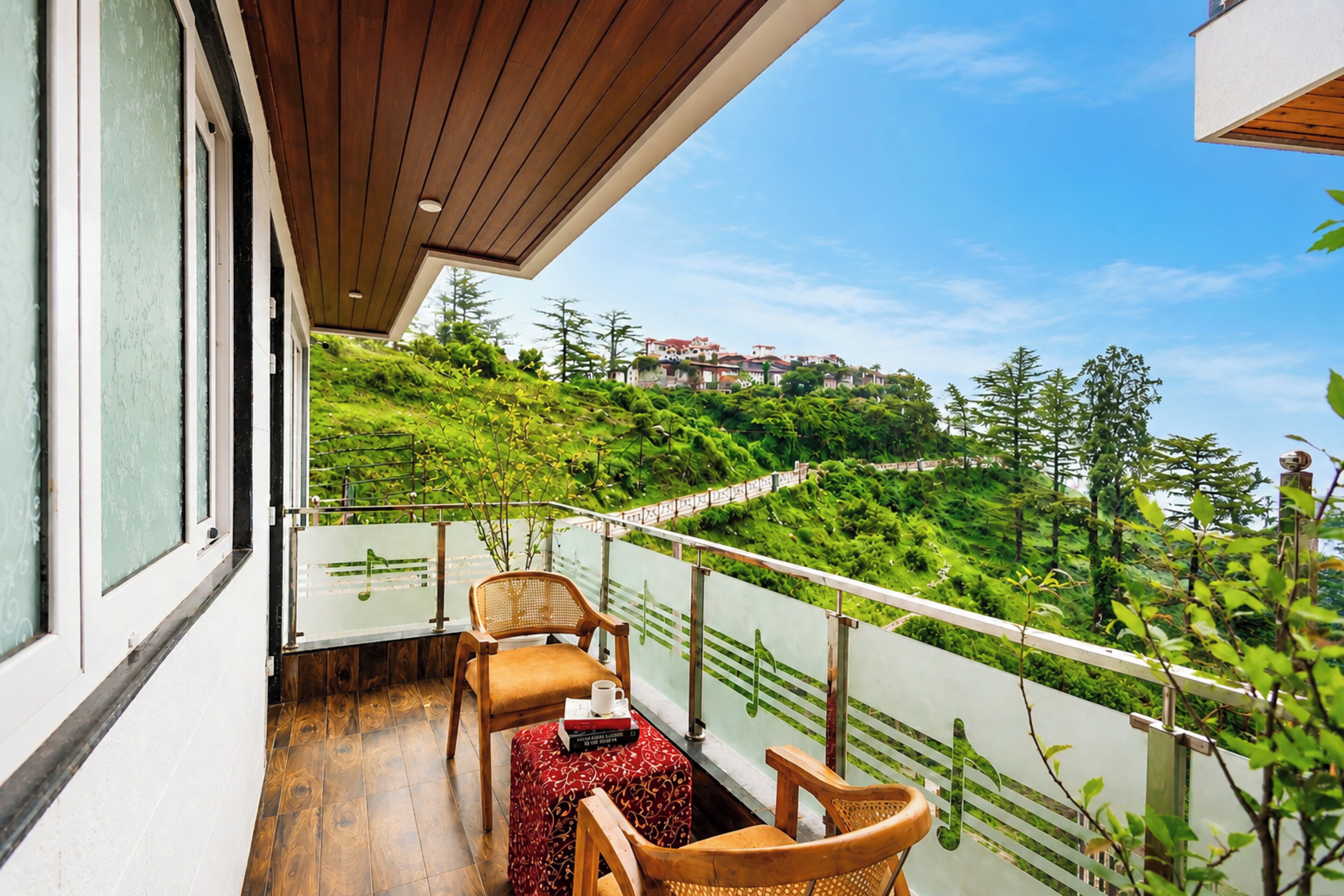 A balcony with a wooden floor, two wicker chairs, a small table with a red patterned tablecloth, and a view of a green hillside with trees and buildings.