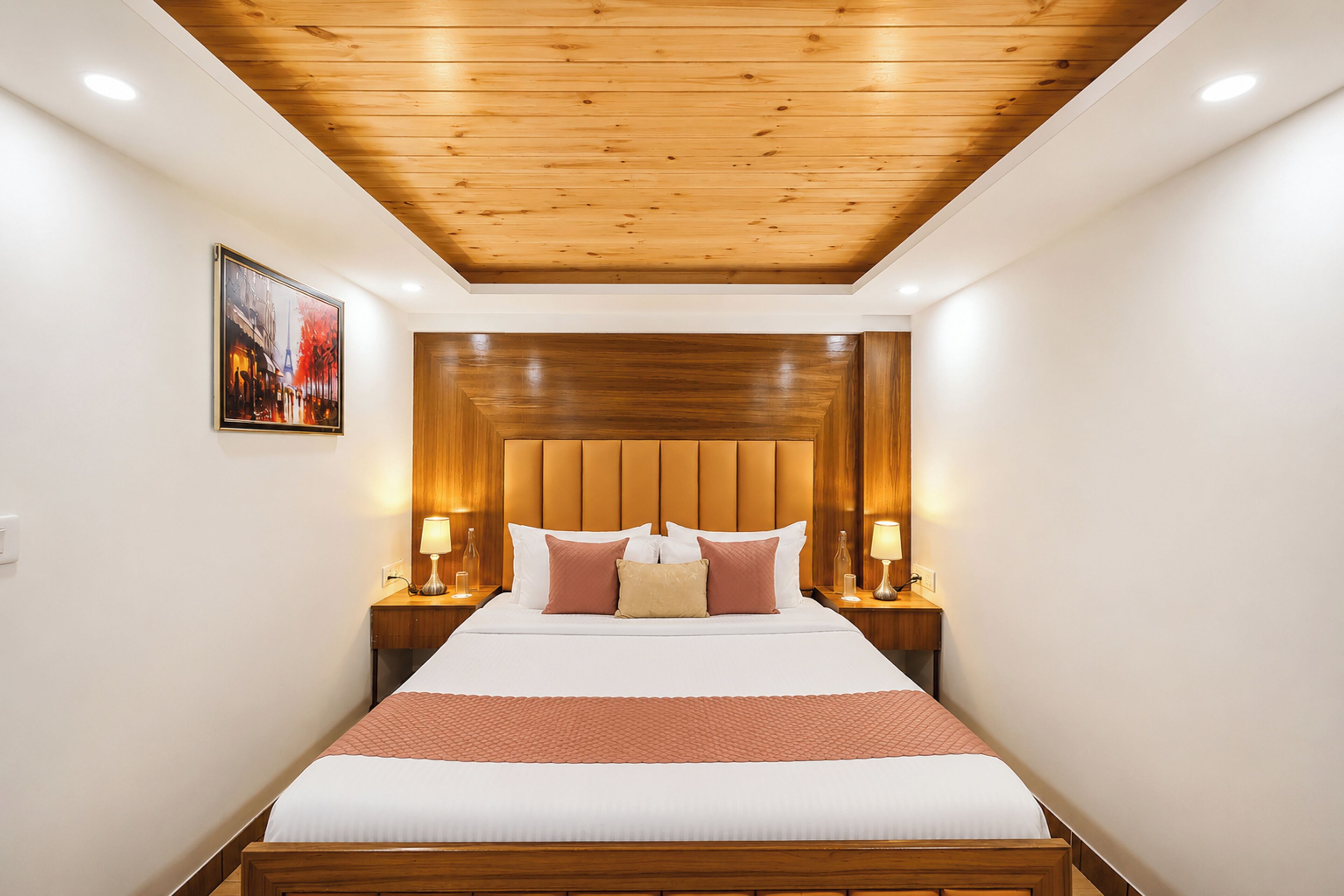 A hotel room with a wooden ceiling, a bed with white bedding and a red accent, two bedside lamps, and a framed picture on the wall.