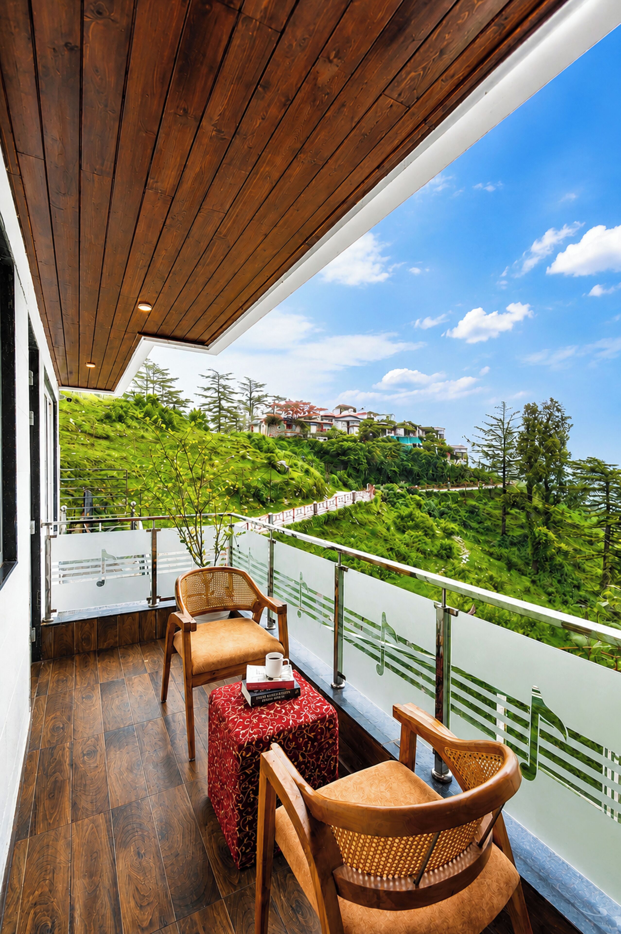 A balcony with wooden flooring, a table with a red patterned tablecloth, two wooden chairs, and a view of a green landscape with buildings in the distance.