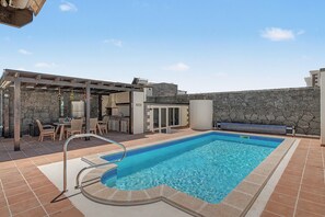 A swimming pool with a stone wall, a covered seating area, and a barbecue setup. - Vacation Villa 'Eire' with Mountain View, Wi-Fi and Air Conditioning (Yaiza)