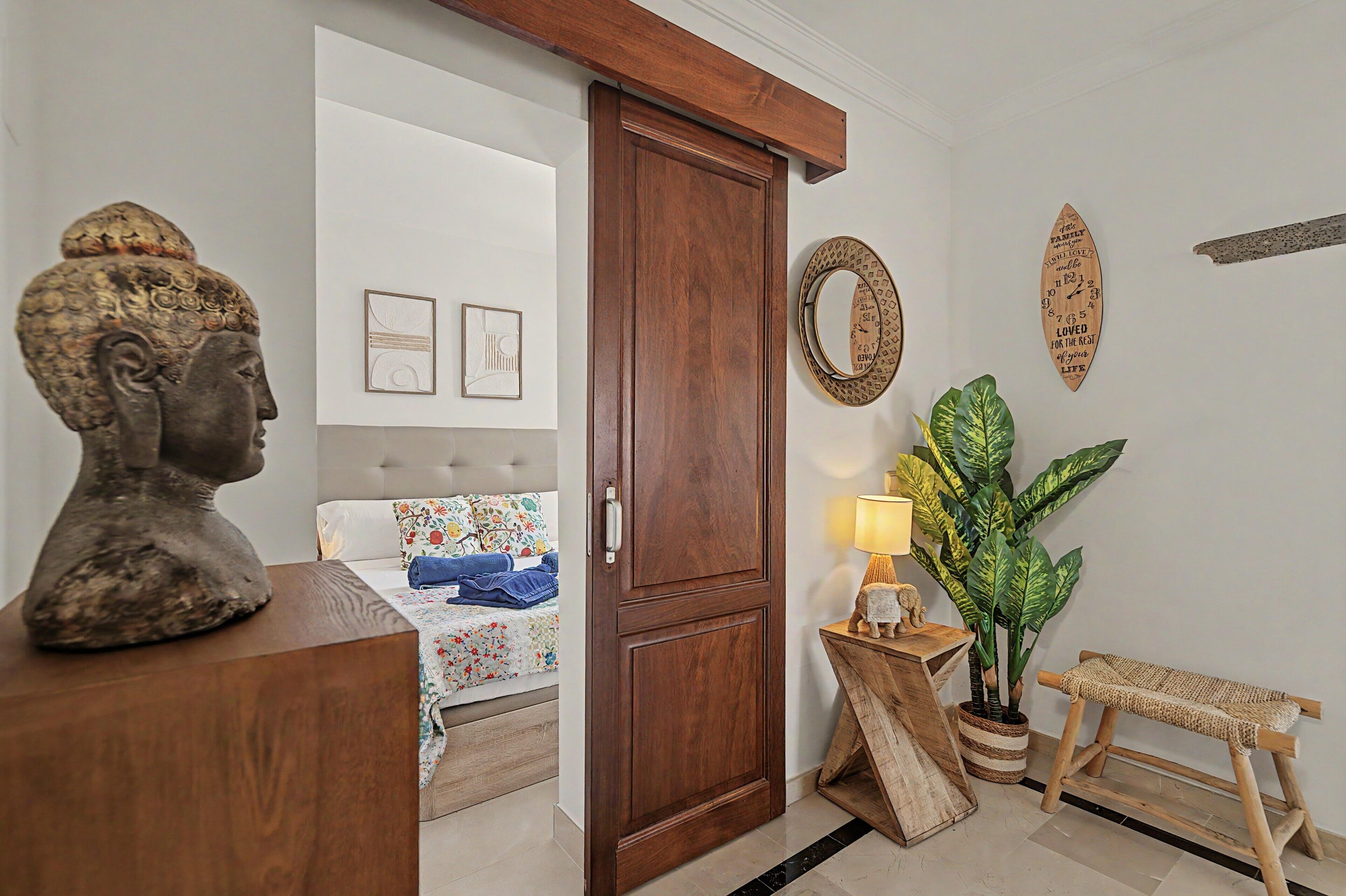 A room with a wooden door, a bust sculpture, a bed with floral bedding, a mirror, and a potted plant.