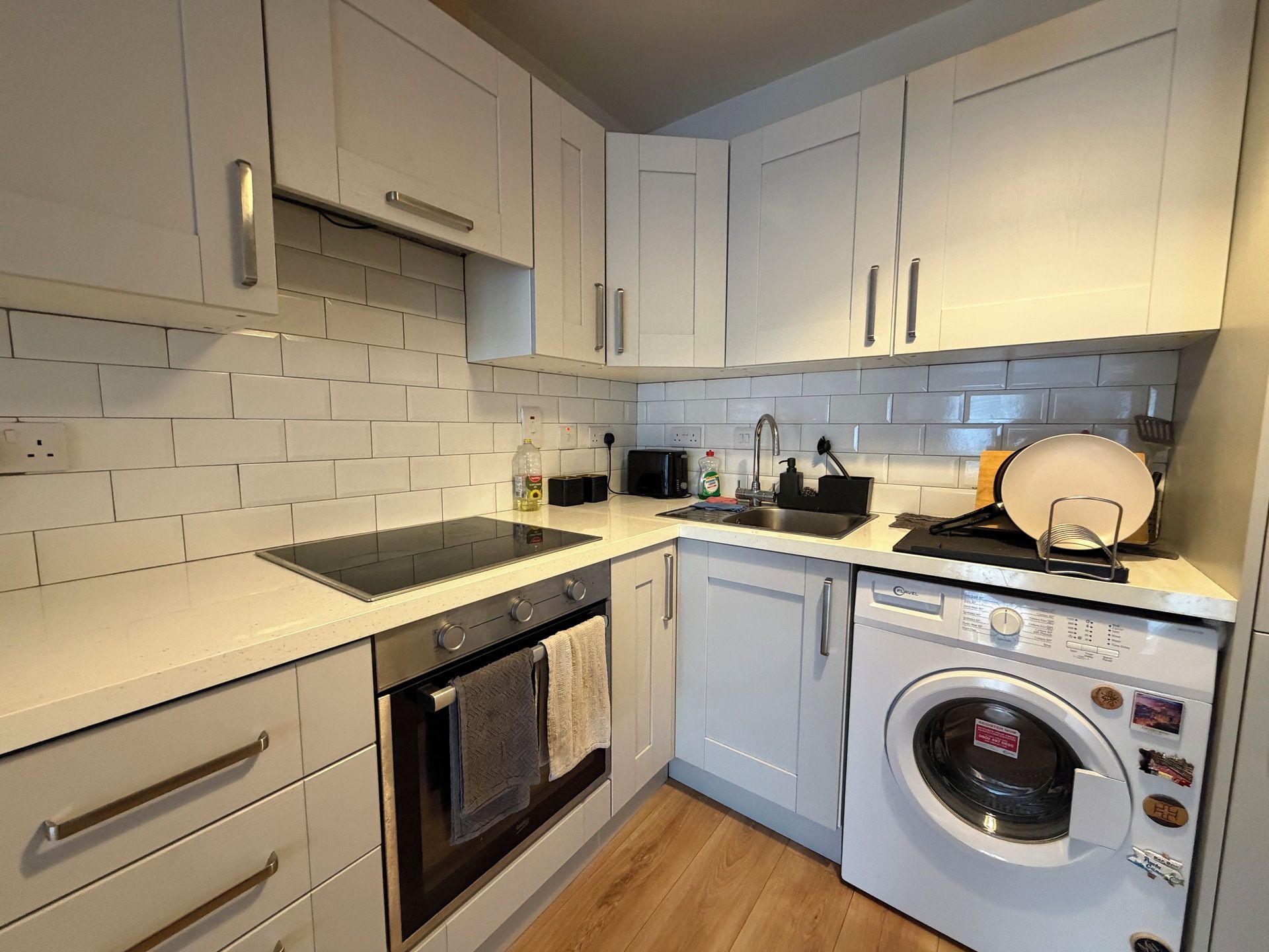 Cocina compacta con armarios blancos, horno integrado, microondas, lavaplatos y lavadora.