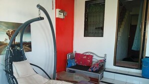 Una habitación con una silla colgante, una mesita, una cama con almohada roja y blanca, y una ventana con reja de metal.