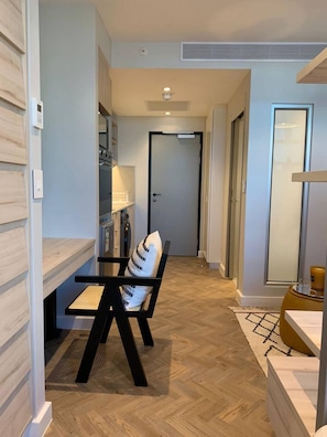 Un interior moderno con piso de madera en patrón espiga, un área de comedor con una silla negra y un cojín blanco, y una cocina con electrodomésticos integrados.