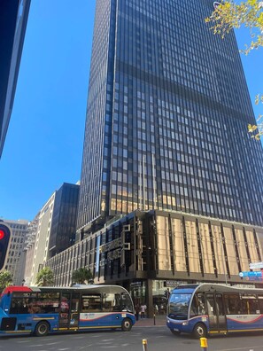 Ein hohes Gebäude mit moderner Glasfassade, ein Geschäft mit der Aufschrift 'FORESTORE PLACE' und zwei vor dem Haus geparkte Busse.