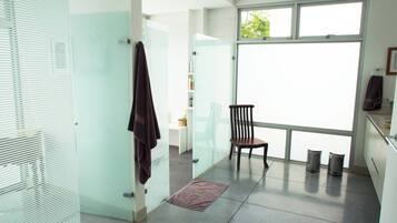 A modern bathroom with a glass shower enclosure, a wooden chair, and a large window providing natural light.