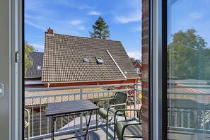 Un balcon avec une table et des chaises, donnant sur un quartier résidentiel avec une maison à toit de tuiles et un ciel dégagé.