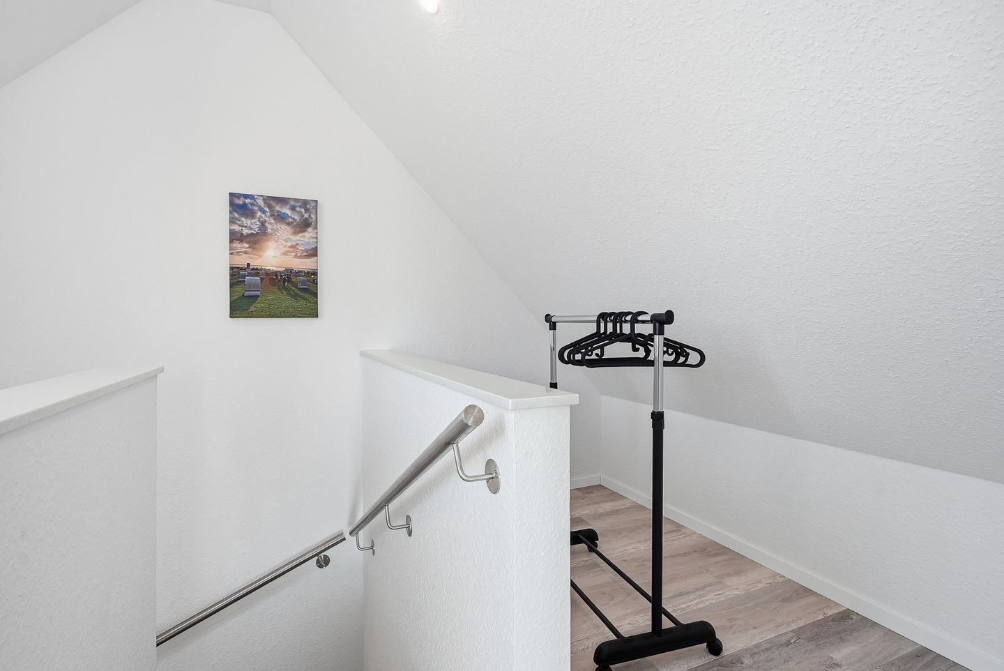 Un coin de pièce avec un mur blanc, un tableau représentant un paysage et un étendoir noir sur lequel sont suspendus des vêtements.