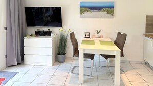 Ein gemütlicher Wohnraum mit Flachbildfernseher, einem Esstisch mit Stühlen, einer kleinen Kommode mit elektronischen Geräten, einer Topfpflanze und einem gerahmten Bild an der Wand.