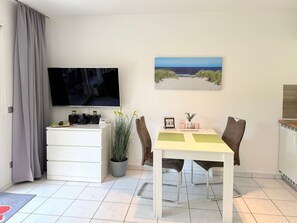 A compact living space with a flat-screen TV, a dining table with chairs, a small cabinet with electronic devices, a potted plant, and a framed picture on the wall. - House at Badeweg 4 - "Sandkorn 1" Apartment (Scharbeutz)