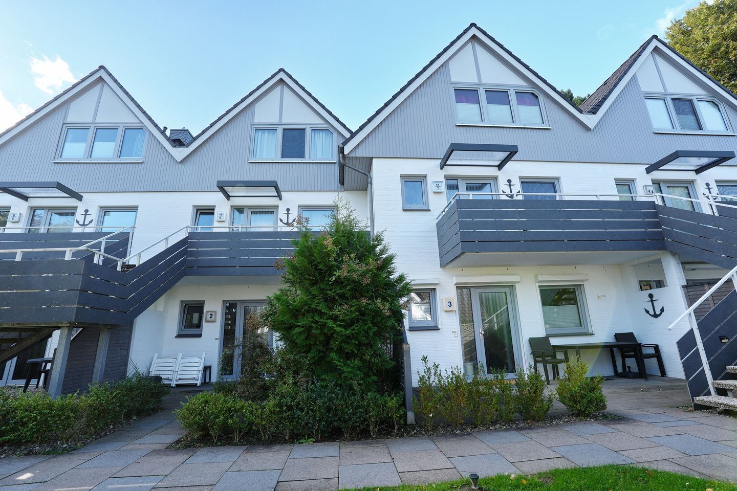 A row of modern, two-story townhouses with balconies, a small tree in the center, and a clear sky.