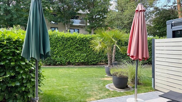 Eine Terrassenfläche mit zwei Sonnenschirmen, einem Rasen, einem Hecken und einem Gebäude im Hintergrund.