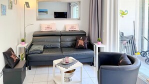 Ein modernes Wohnzimmer mit einer Couch, einem Couchtisch und einem Sessel. Es gibt ein großes Fenster mit Vorhängen, einen Spiegel über der Couch und einen kleinen Tisch mit einer Pflanze.