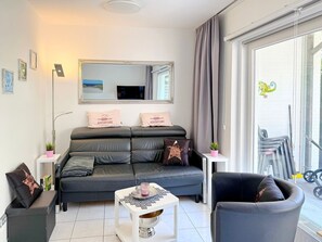 A modern living room with a sofa, a coffee table, and a chair. There is a large window with curtains, a mirror above the sofa, and a small table with a plant. - House at Badeweg 4 - "Sandkorn 1" Apartment (Scharbeutz)