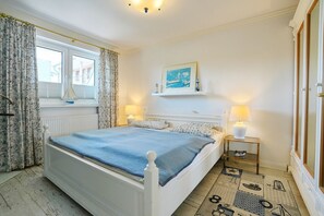 A bedroom with a bed, two bedside lamps, a window with curtains, a wall-mounted shelf with a picture, and a patterned rug. - Seebrücke Apartment 11 - Seebrücke (Scharbeutz)