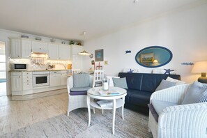 Eine moderne Küche mit weißen Schränken, ein Essbereich mit rundem Tisch und Korbstühlen sowie ein Wohnzimmer mit einem dunklen Sofa und gestreiften Kissen.