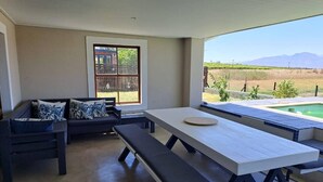A covered patio area with a dining table and benches, a sofa with cushions, and a view of a swimming pool and landscape. - Country Retreat with Pool & Braai, Sleeps 6 (Riebeek-Kasteel)
