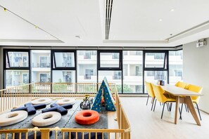 Een moderne kamer met grote ramen, een speelruimte met kussens en een tent, en een eetgedeelte met gele stoelen.