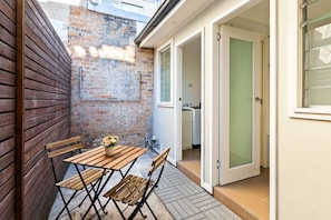 Eine kleine Terrasse mit Holztisch und -stühlen, einer Ziegelwand und einer Tür zu einem Zimmer.