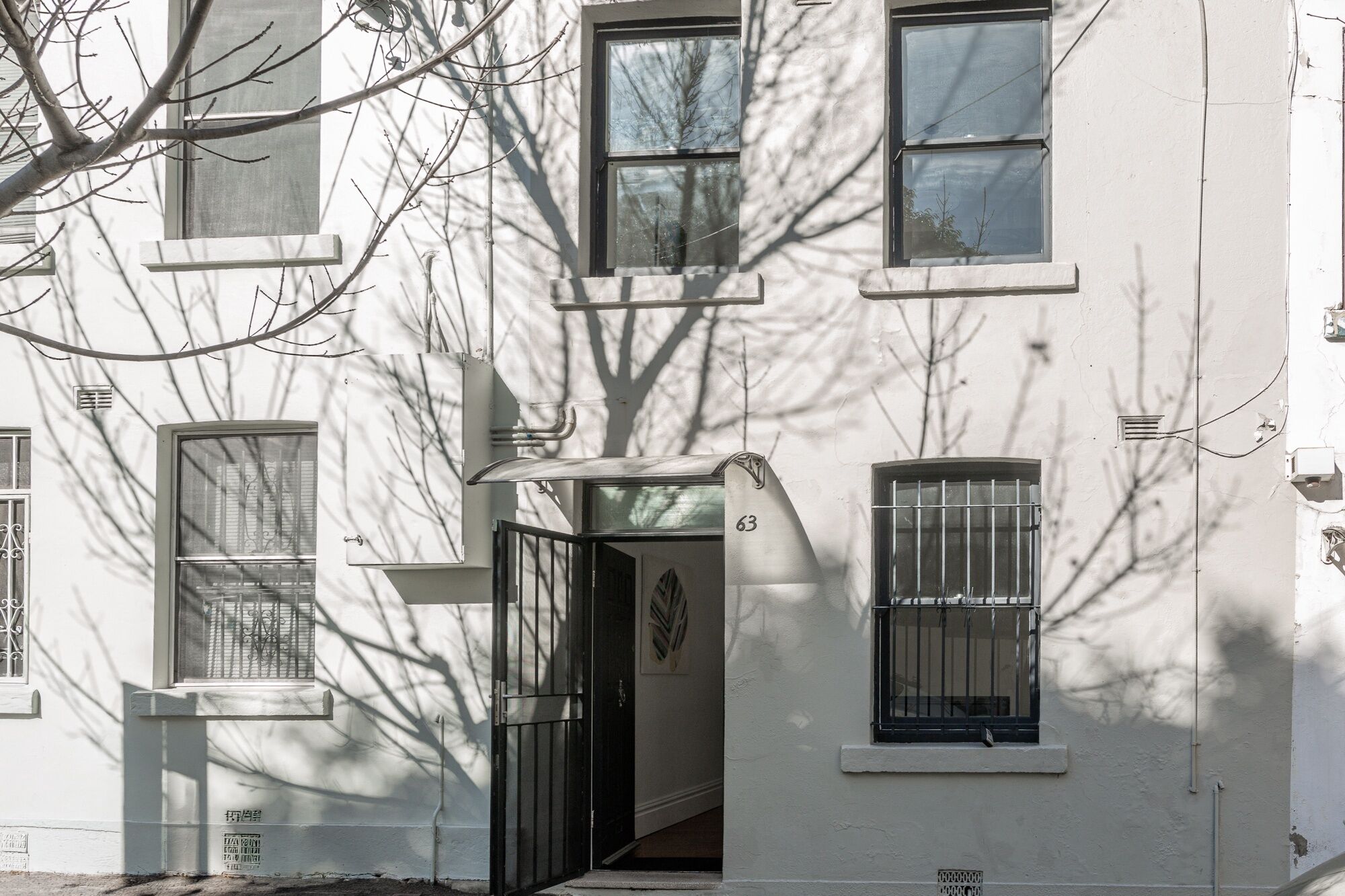 A white building with multiple windows, a door with a number '63' sign, and a tree casting shadows on the facade.