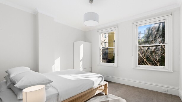 Ein Schlafzimmer mit einem Bett, einem Nachttisch, einer Lampe und zwei Fenstern mit Blick auf Gebäude und Bäume.