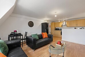 Ein moderner Wohnbereich mit einem schwarzen Ledersofa, einem Glastisch und einer Küchenzeile mit Holzschränken und einem schwarzen Kühlschrank.