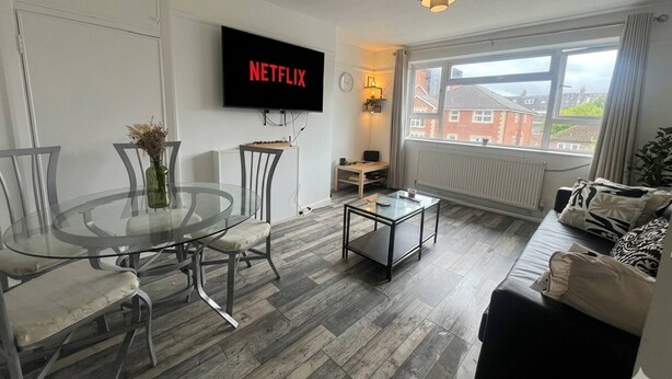 A modern living room with a glass dining table, a sofa, a coffee table, and a flat-screen TV displaying 'NETFLIX'. - Ultra-Spacious 2 BR In Central Wimbledon! (Greater London)