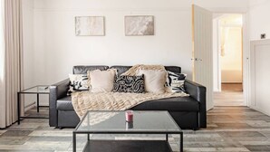 A modern living room with a black leather sofa, two framed abstract paintings, a glass coffee table, and a door leading to another room. - Ultra-Spacious 2 BR In Central Wimbledon! (Greater London)