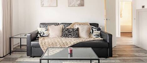 A modern living room with a black leather sofa, two framed abstract paintings, a glass coffee table, and a door leading to another room.