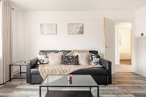 Ein modernes Wohnzimmer mit einem schwarzen Ledersofa, zwei gerahmten abstrakten Gemälden, einem Glastisch und einer Tür zu einem anderen Raum.