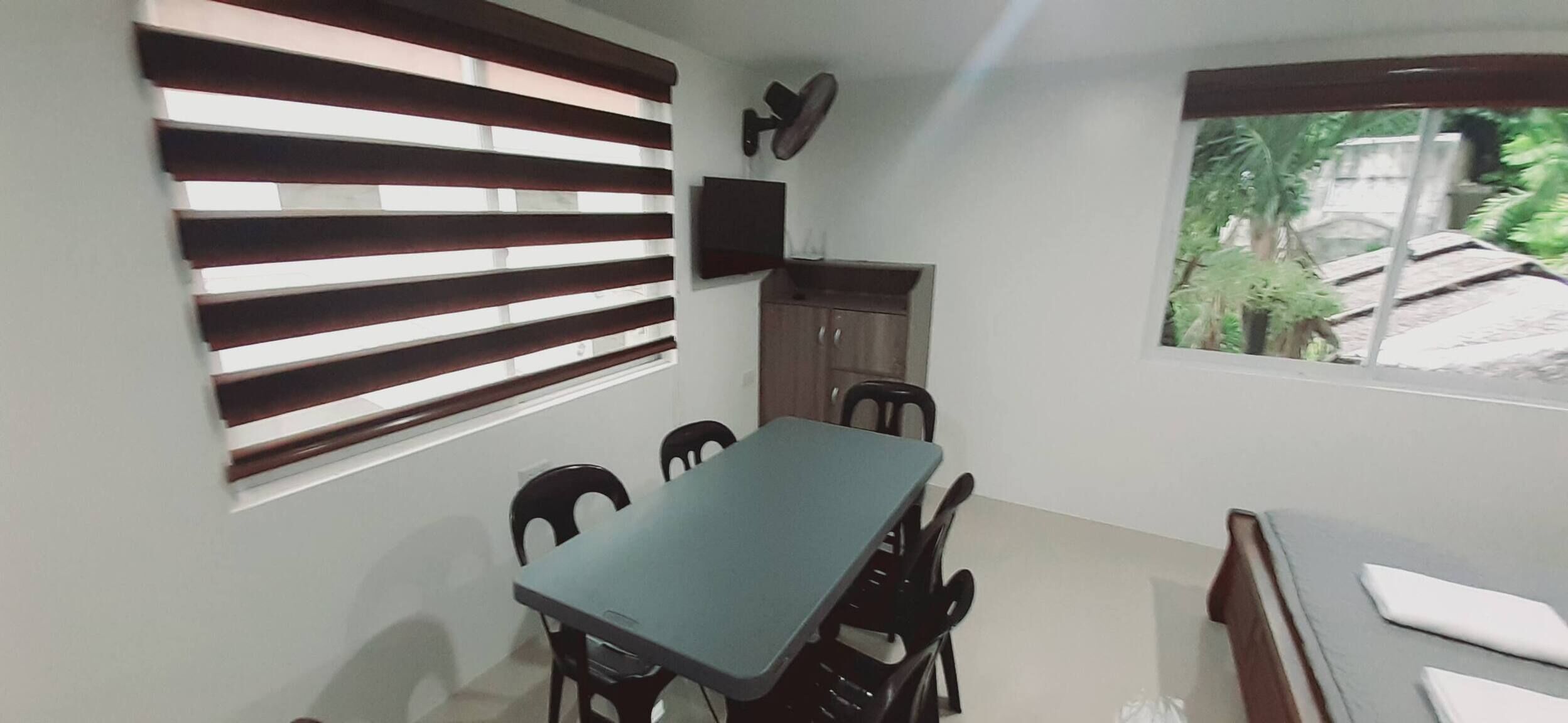 A dining area with a table and chairs, a television on the wall, and a window with a view of greenery.