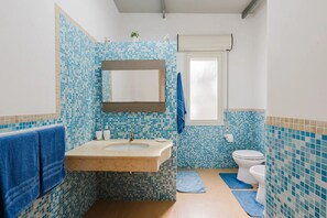 A bathroom with a tiled mosaic backsplash, a wooden vanity, a sink, a mirror, a toilet, and blue towels. - Charming Villa Rental in Mazara Del Vallo with Pool Access (Mazara Del Vallo)