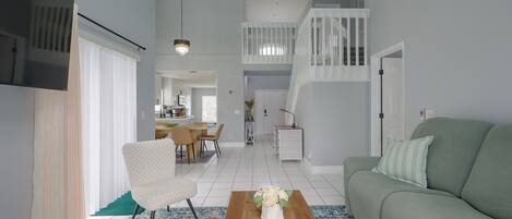 A modern living room with a green sofa, a white armchair, and a wooden coffee table. There is a staircase leading to an upper level, and a kitchen area is visible in the background.