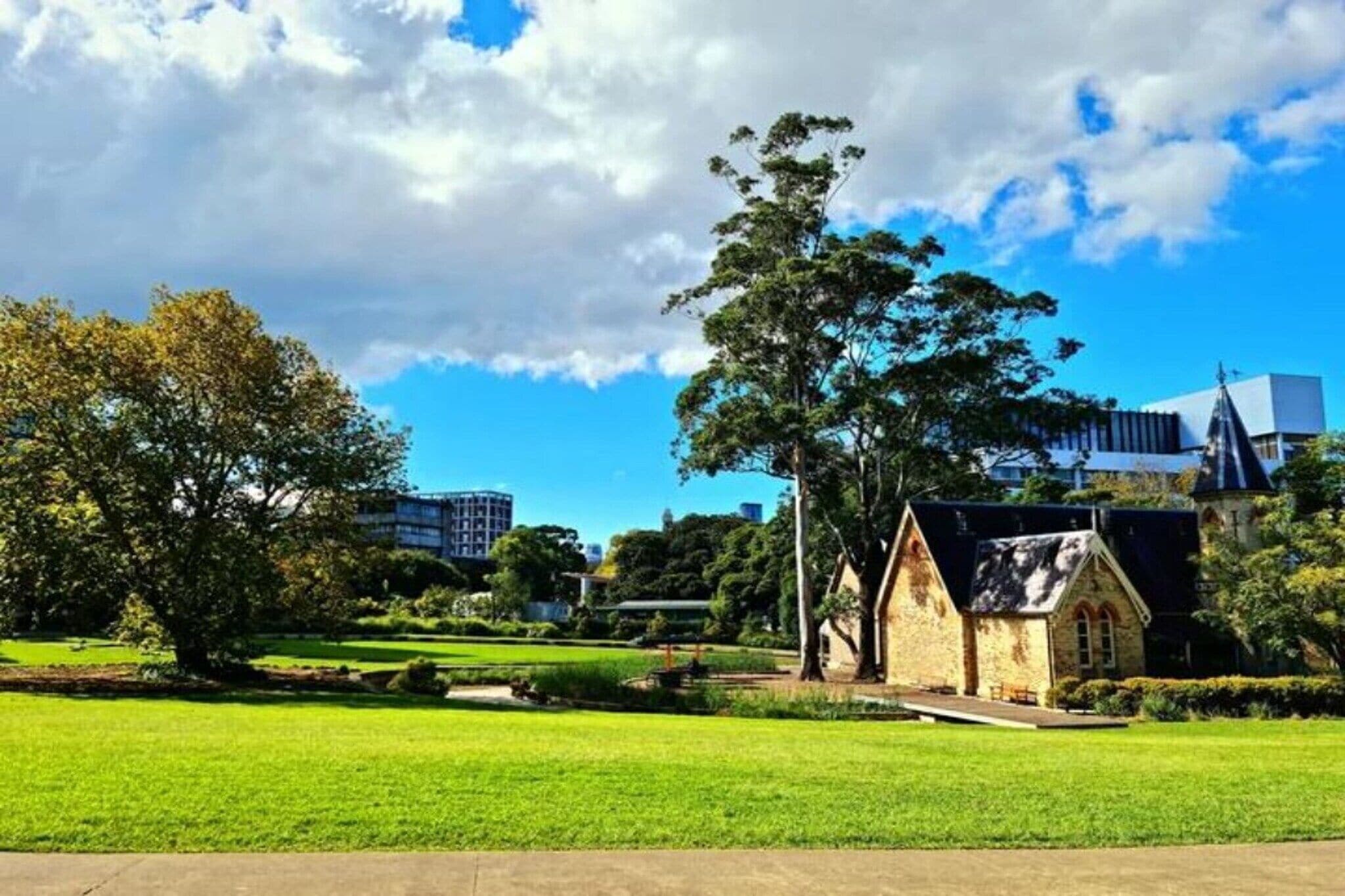 A park with a small stone building, a large tree, and modern buildings in the background.