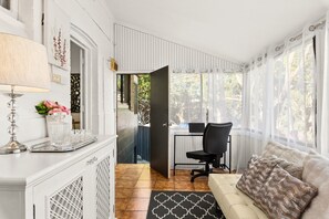 Ein Zimmer mit einem weißen Schrank, einem Schreibtisch mit Stuhl, einer Couch mit gemusterten Kissen und Blick durch ein Fenster mit transparenten Vorhängen.