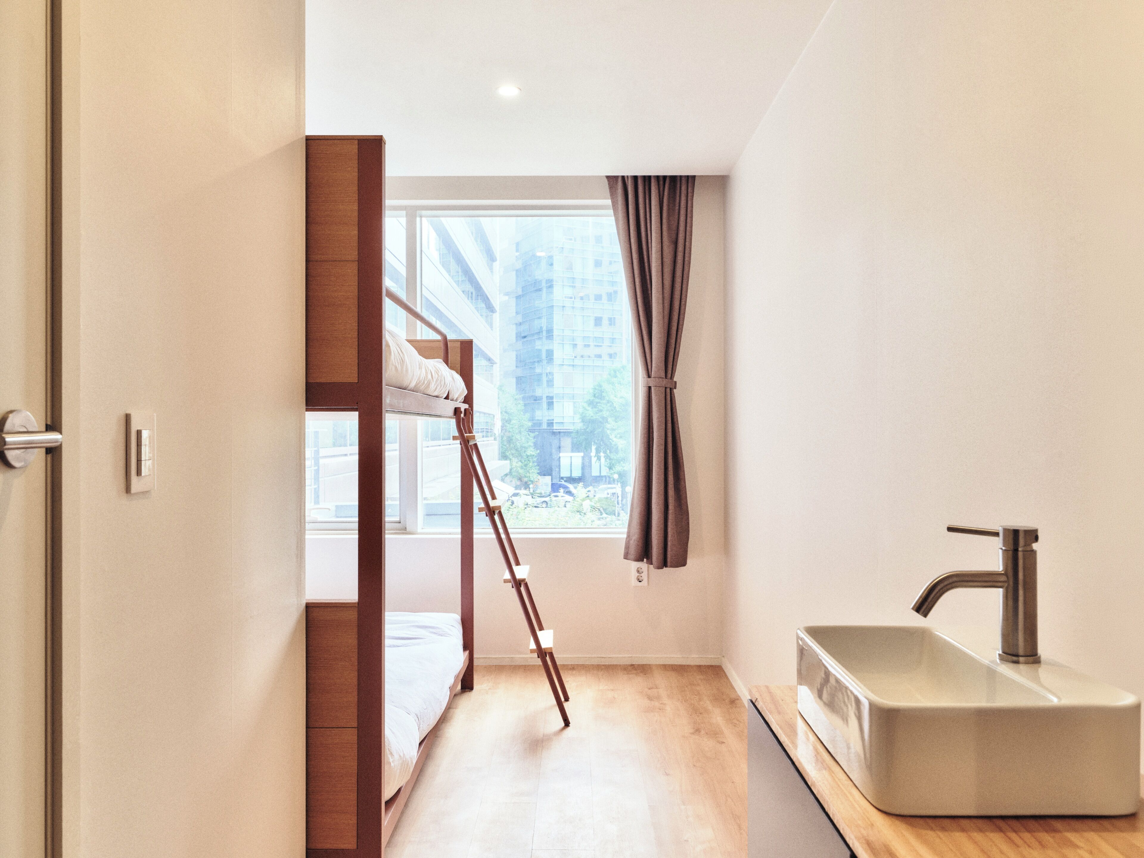 Une chambre à coucher de taille modeste, avec un lit superposé, une petite fenêtre avec des rideaux, un lavabo et une vue sur un bâtiment à l’extérieur.