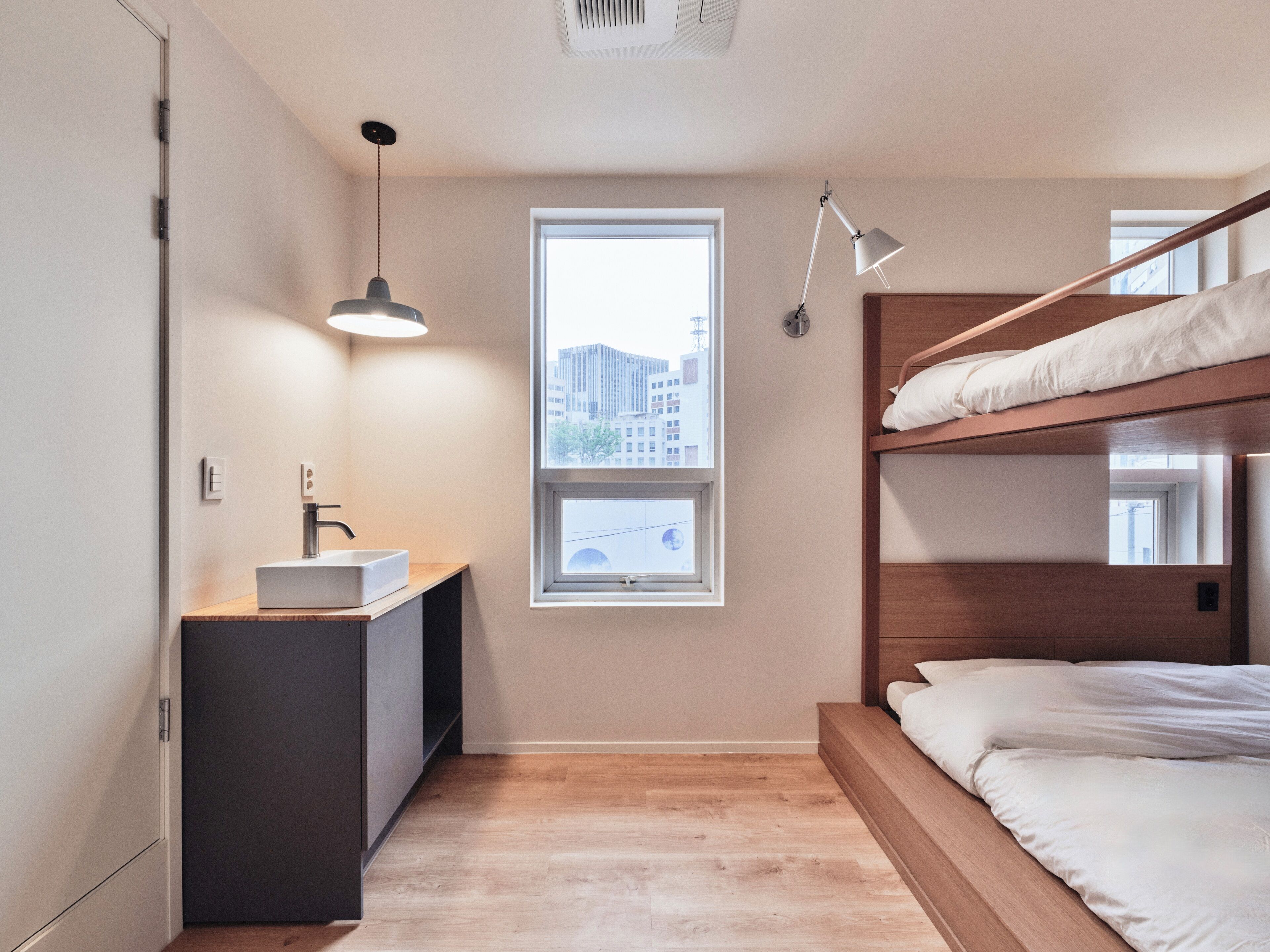 A compact bedroom with a bunk bed, a bedside table with a sink, a window with a city view, and a pendant light.