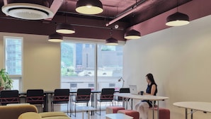 A modern office space with a large window, multiple tables, chairs, and a person working on a laptop. - Susu House (Seoul)