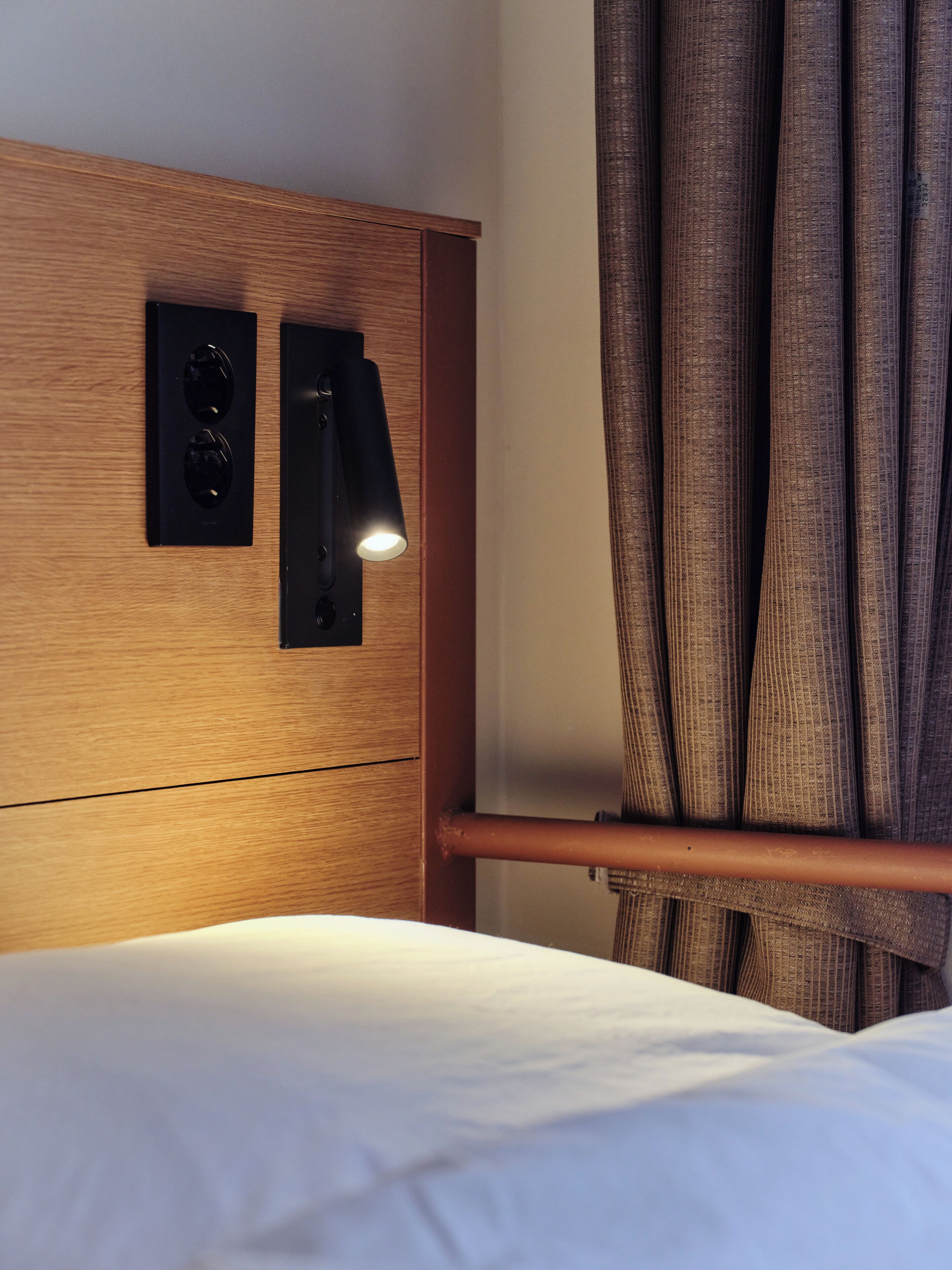Une chambre d’hôtel avec une tête de lit en bois, une table de chevet, une lampe et un rideau.