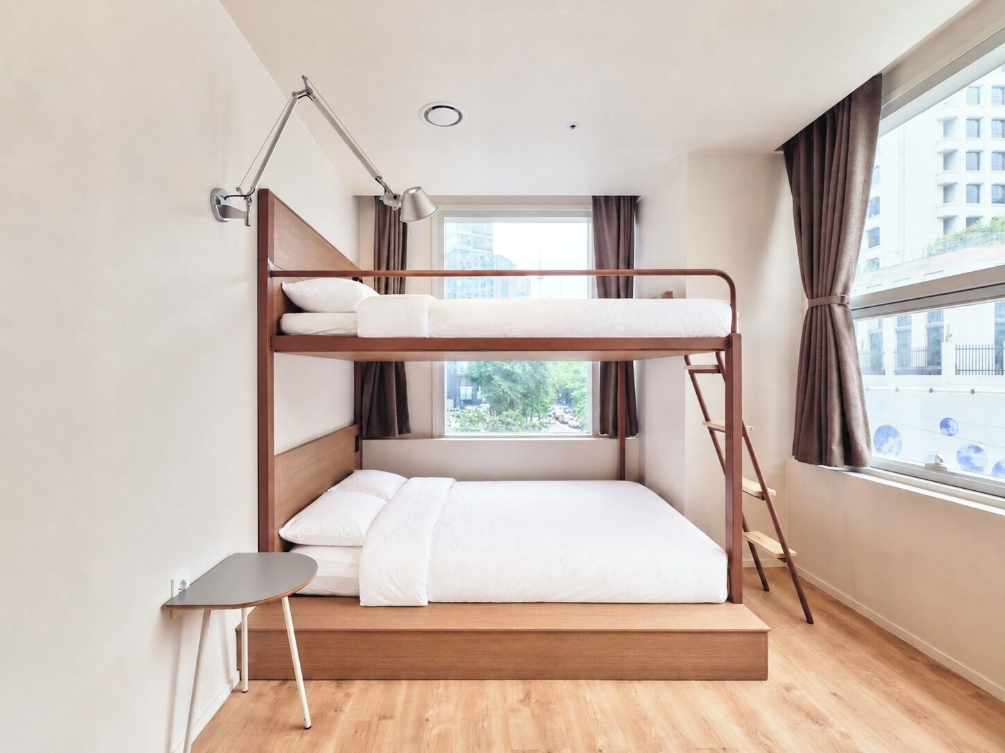 A bunk bed with a desk and lamp, a window with curtains, and a view of buildings outside.