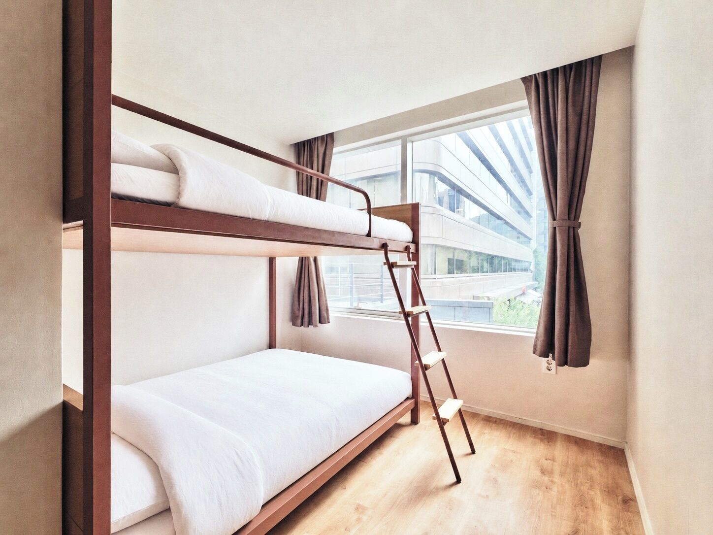 A bunk bed room with a wooden ladder, a window with curtains, and a view of a building outside.